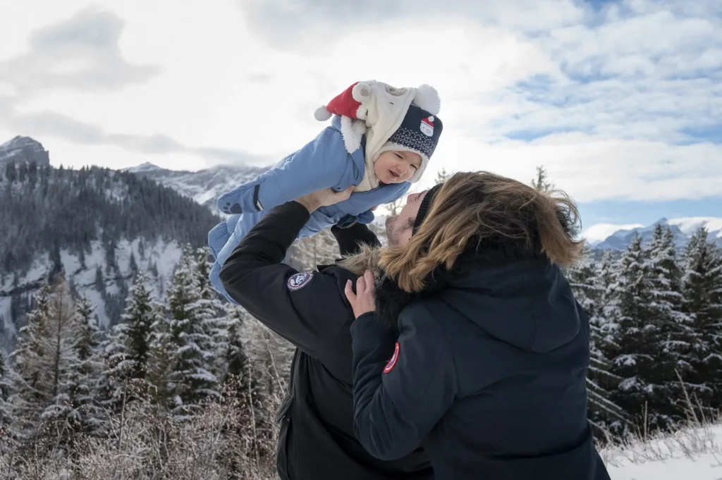 Adulte soulevant un bébé dans ses bras sur fond de paysage enneigé avec sapins et montagnes.
