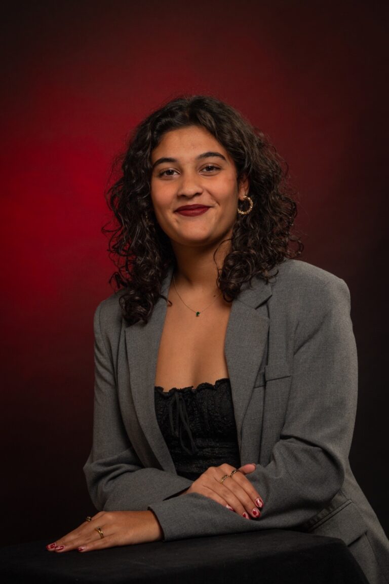 Portrait studio d’une femme en blazer gris sur fond dégradé rouge et noir, assise et souriante.